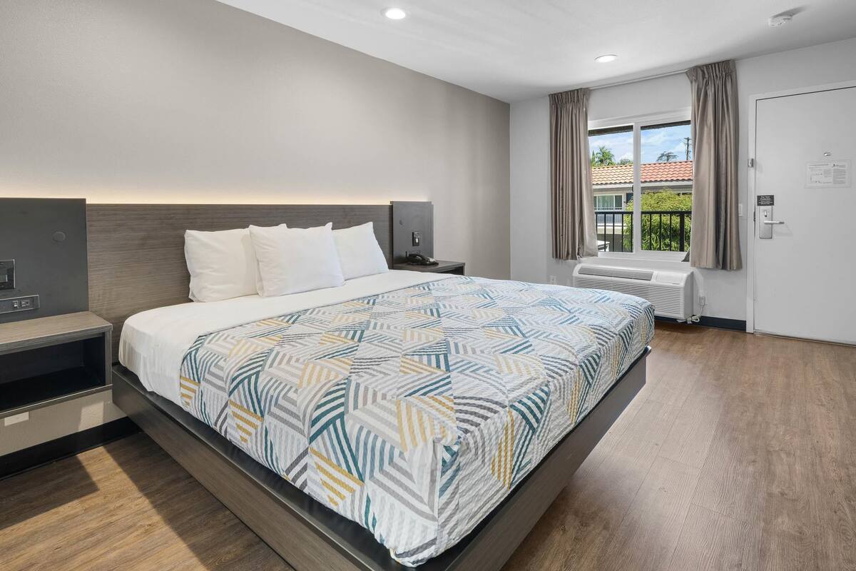 Quarto de hotel moderno com cama grande, mesa de cabeceira, televisão embutida e janela com cortinas.