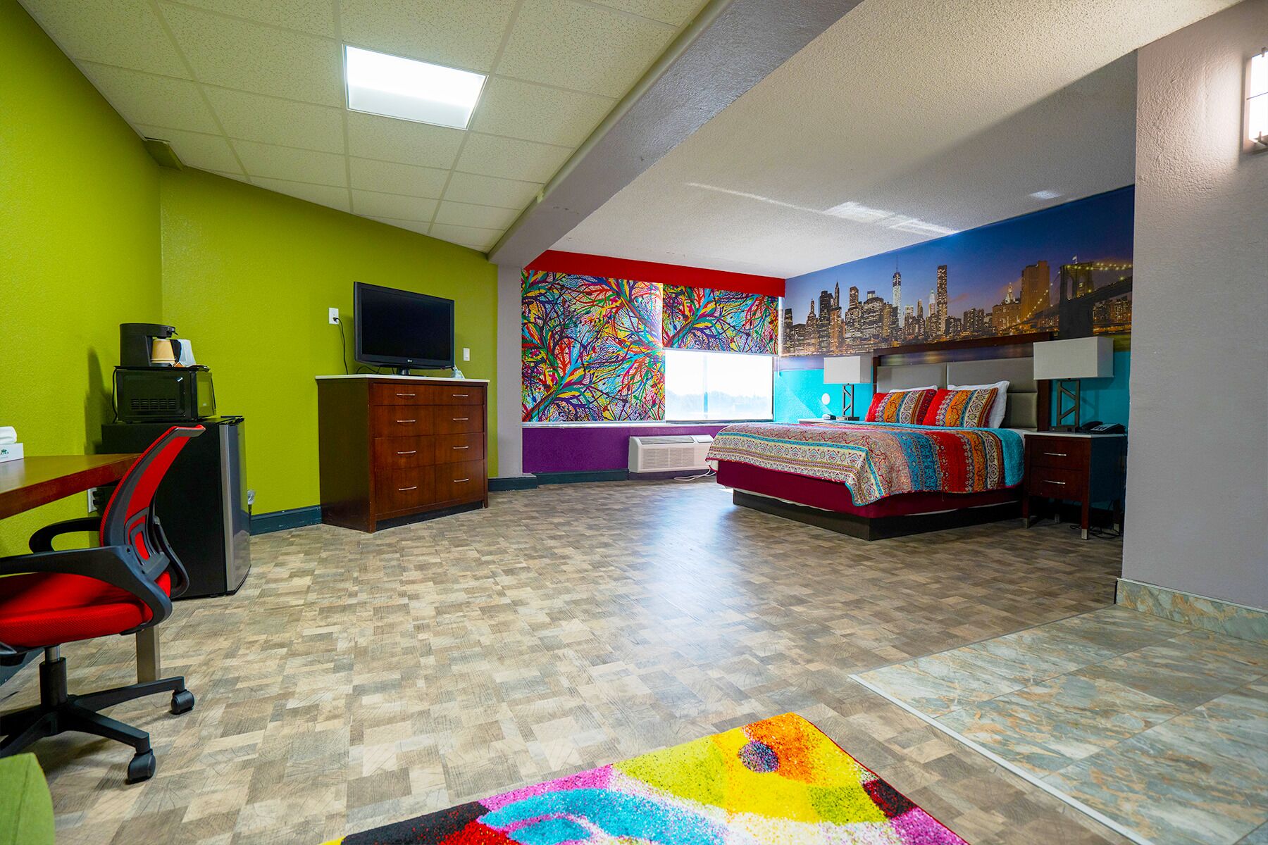A hotel room with a cityscape mural, a bed with a colorful bedspread, a desk with a computer, and a chair.