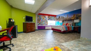 A hotel room with a cityscape mural, a bed with a colorful bedspread, a desk with a computer, and a chair.