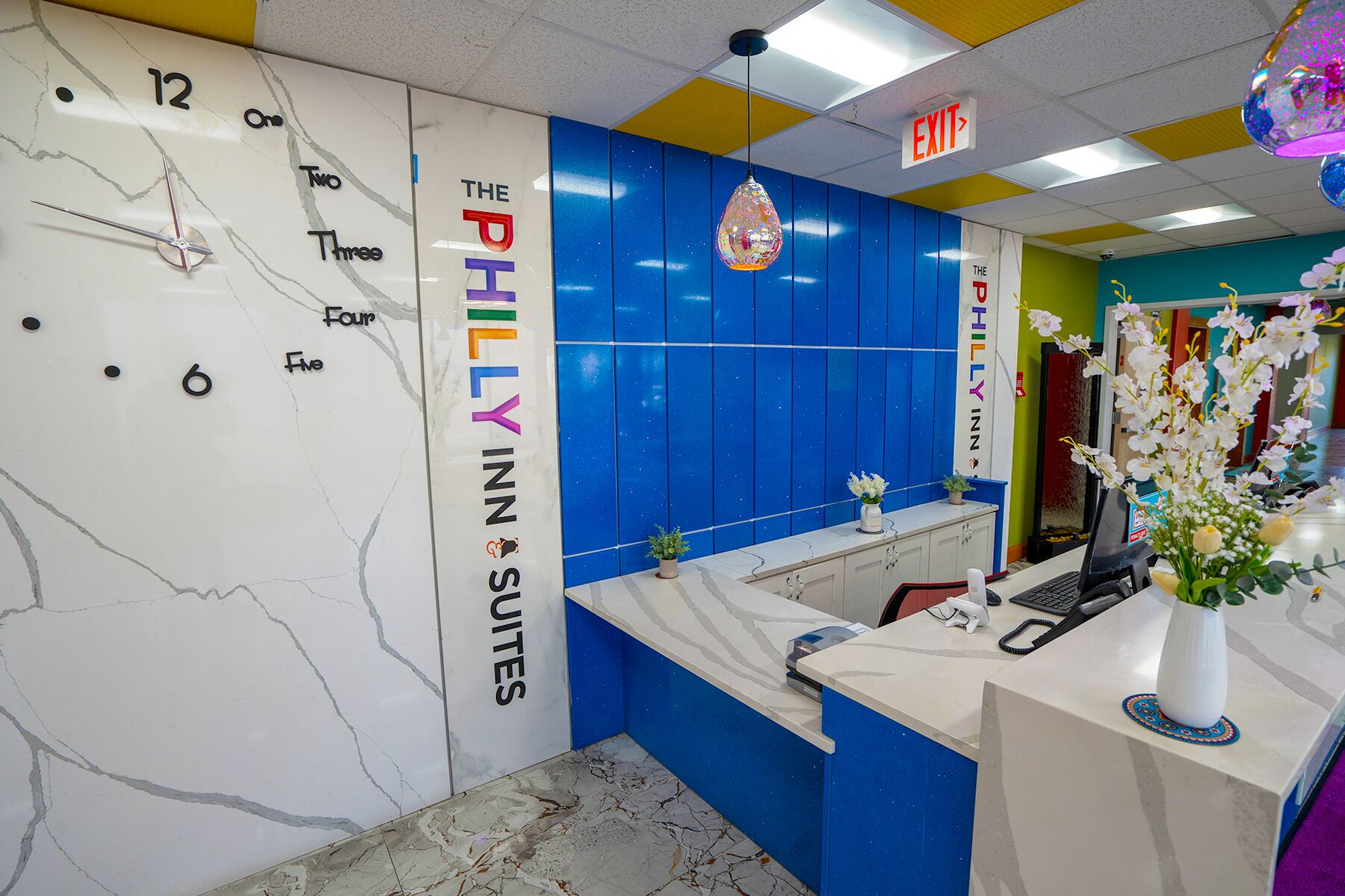 A hotel reception area with a marble wall displaying room numbers, a blue wall with the hotel name, a reception desk with a computer and phone, and a hanging decorative light.