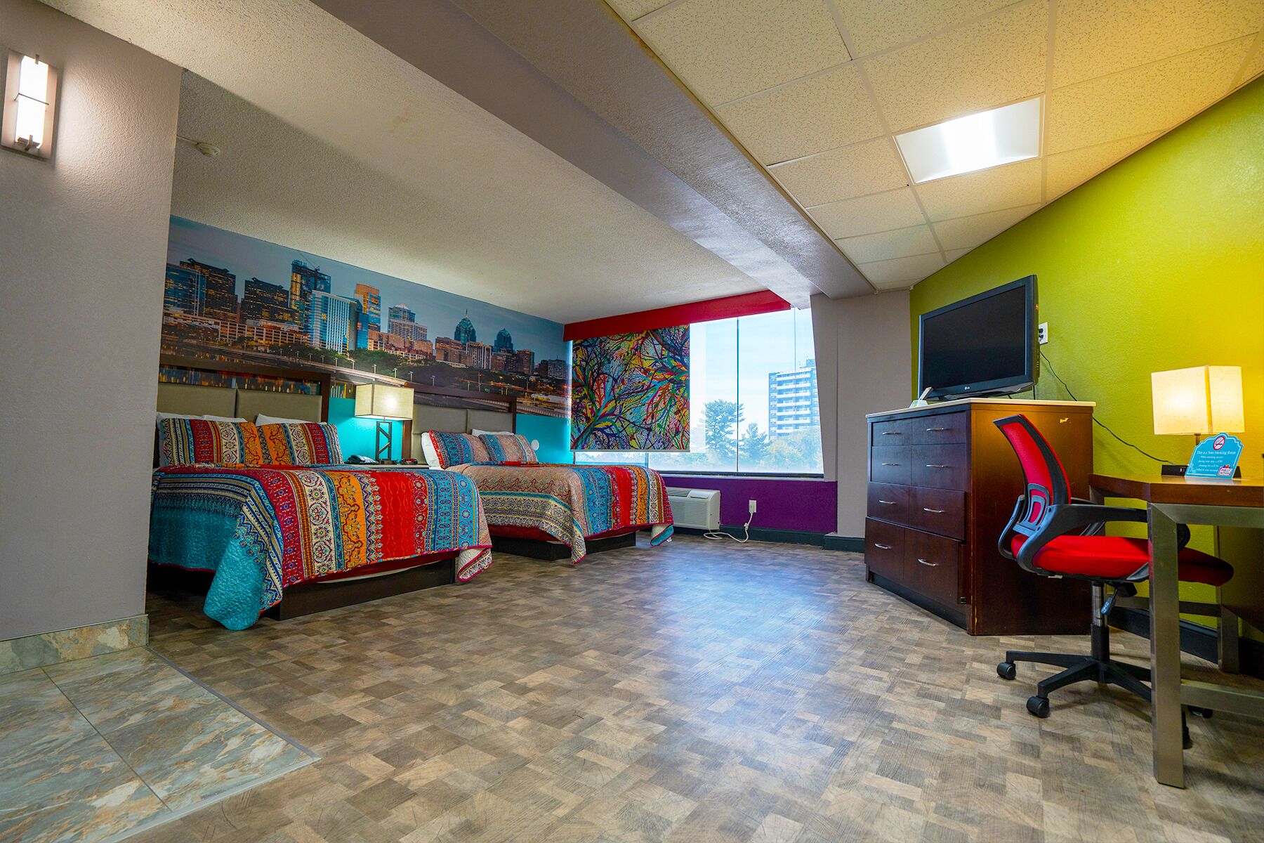A hotel room with two beds, a desk with a computer, a chair, a TV, and a window with a cityscape view.