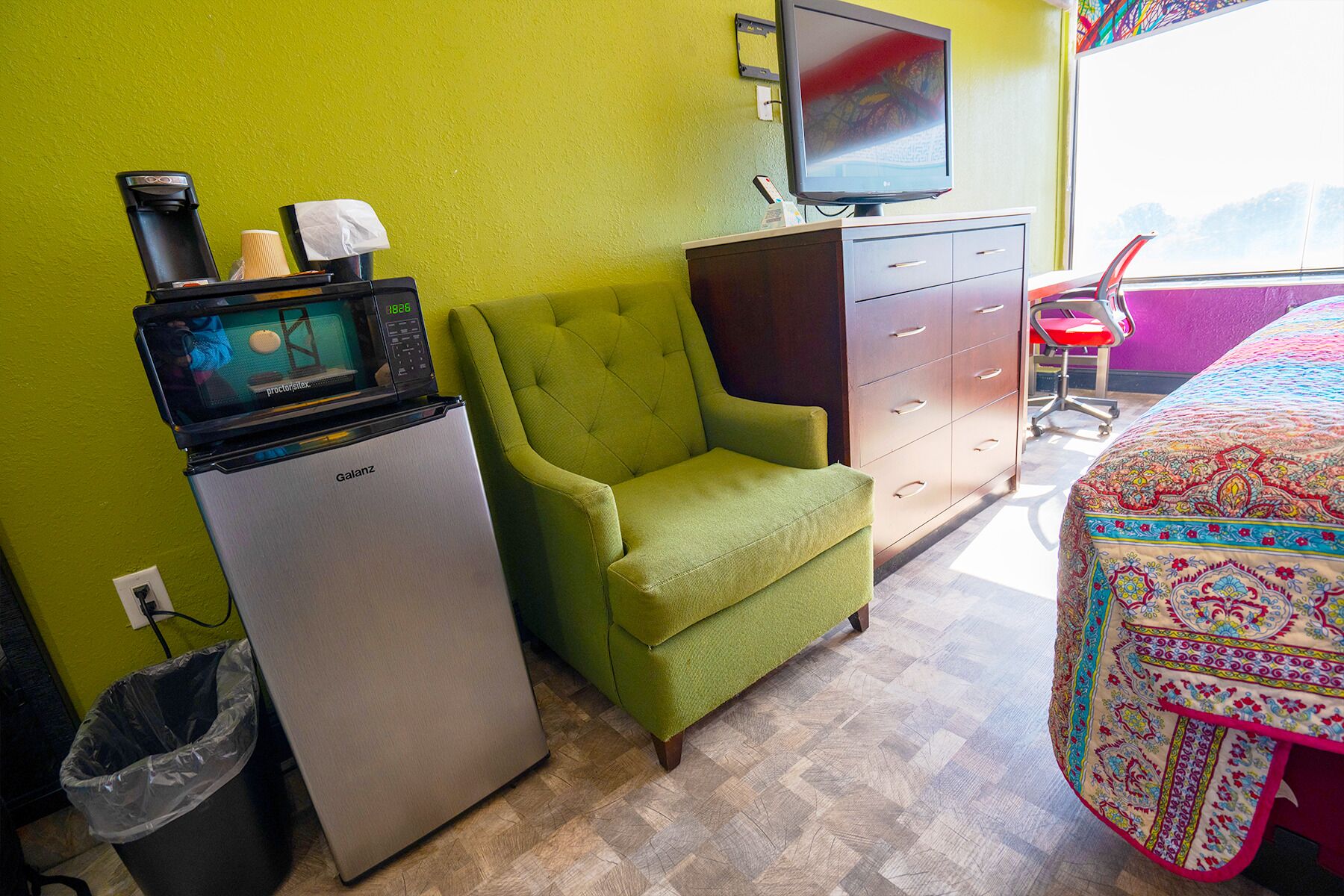 Quarto de hotel pequeno com uma poltrona verde, um frigorífico, uma televisão sobre um suporte, uma secretária com cadeira e um edredom estampado.