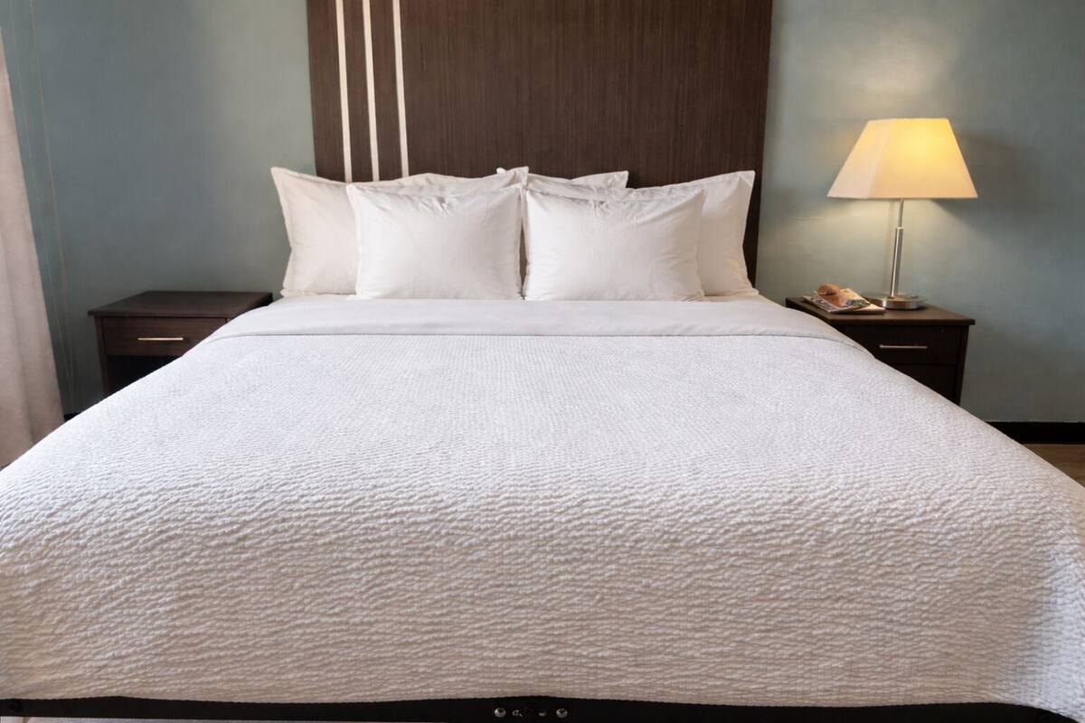 A neatly made bed with white bedding, two pillows, and a bedside lamp on a wooden nightstand.