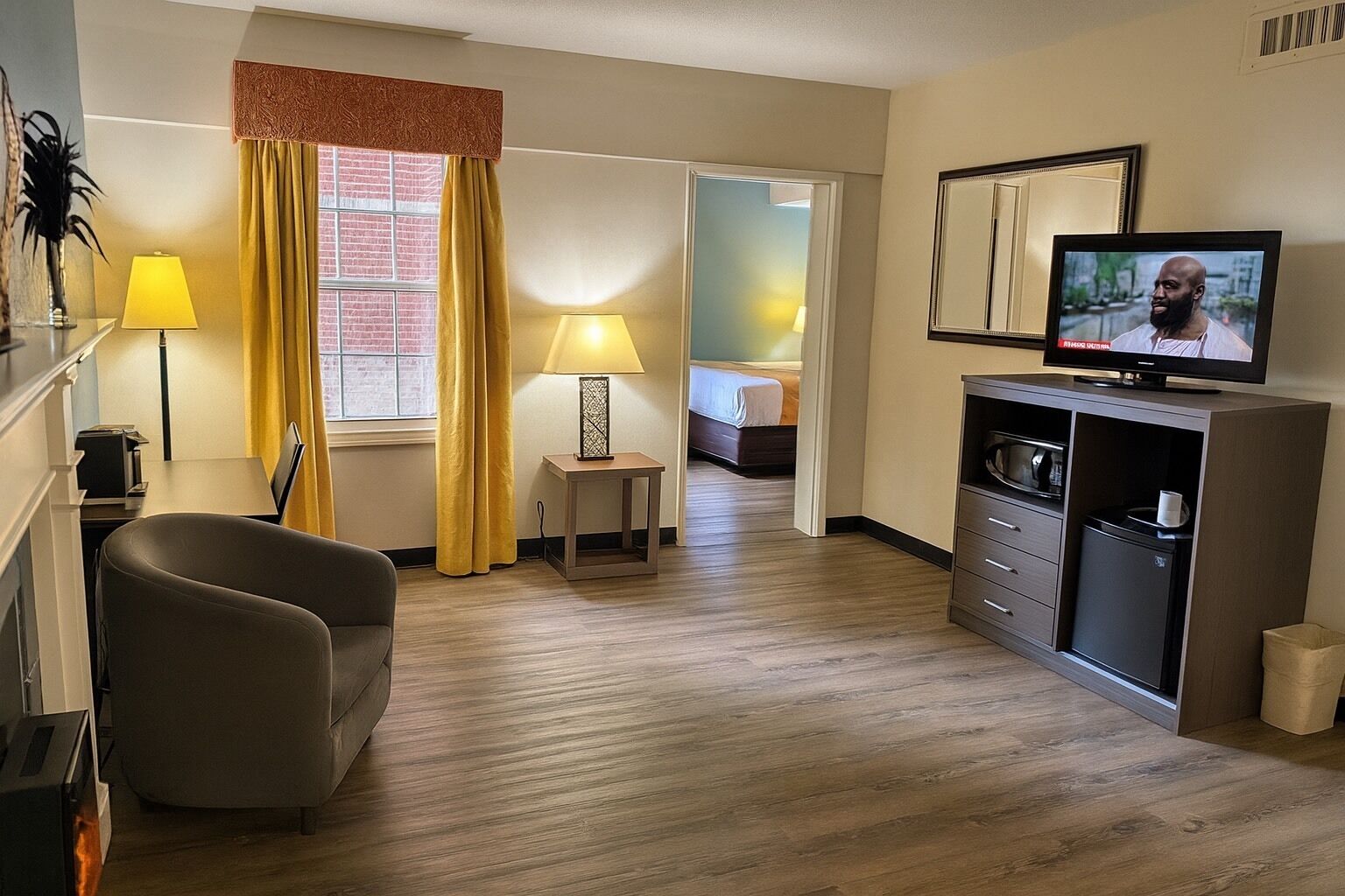A hotel room with a sofa, a TV on a stand, a small table with a lamp, and a bed in the background.