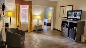 A hotel room with a sofa, a TV on a stand, a small table with a lamp, and a bed in the background.