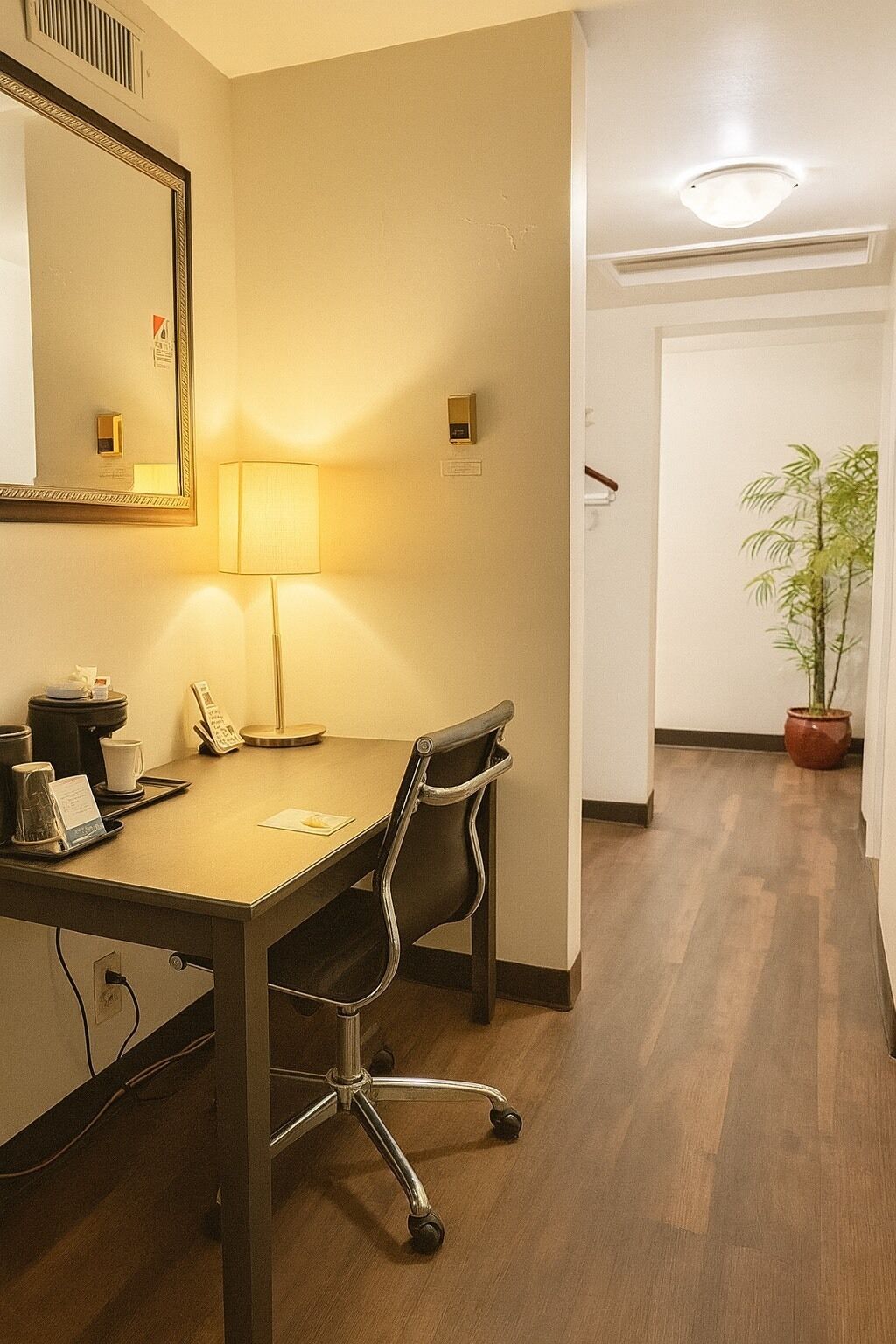 Une chambre d’hôtel avec un bureau en bois, une chaise, une lampe, un miroir, une plante en pot et une porte donnant sur une autre pièce.