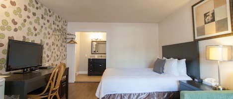Habitación de hotel con cama, escritorio con televisor, silla, mesita de noche con lámpara y baño visible a través de una puerta abierta.