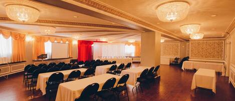 Un salón de conferencias con filas de mesas y sillas, grandes candelabros y un escenario con cortina roja.