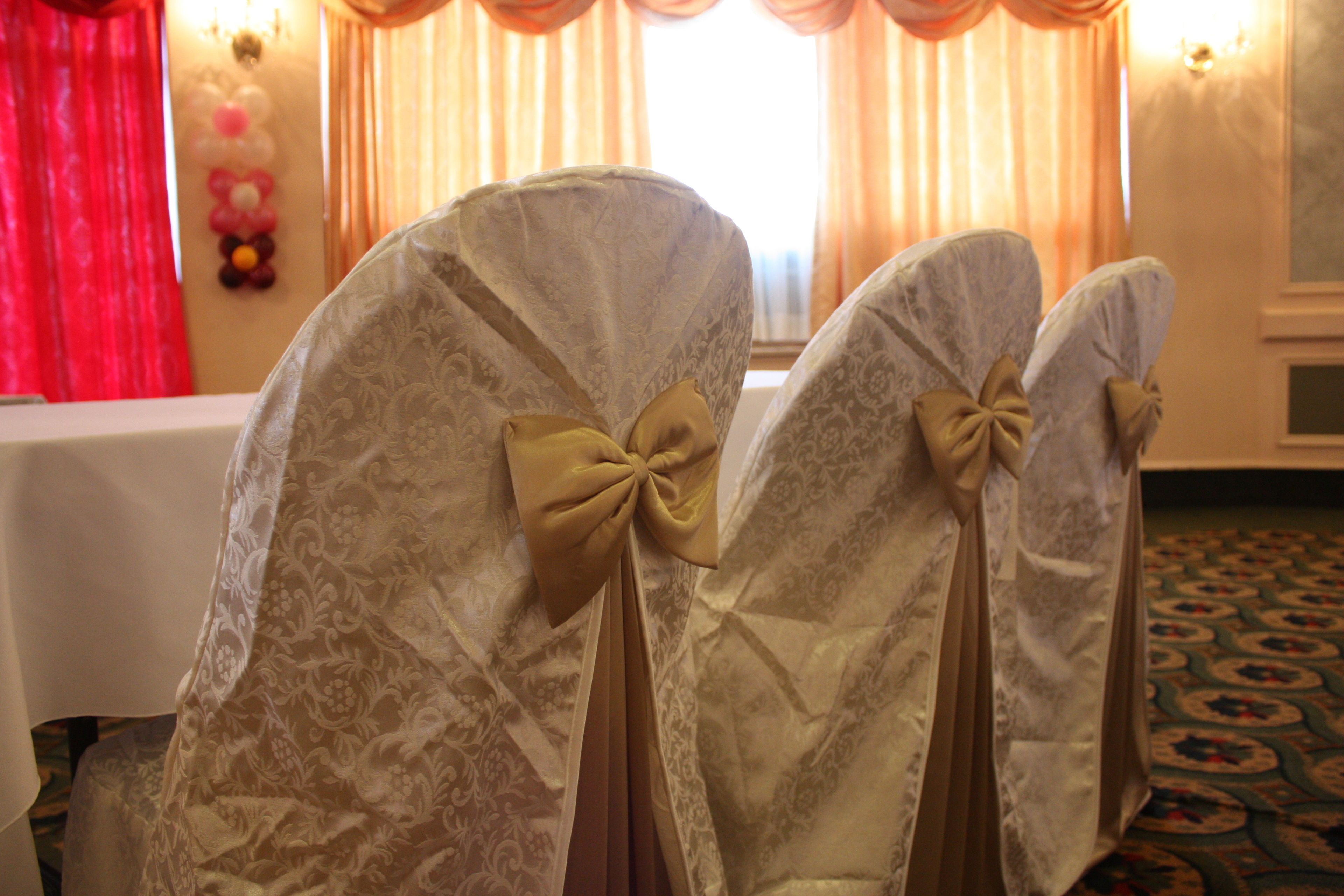 Two chairs with patterned fabric and bow ties, positioned in a room with a table and curtains in the background.