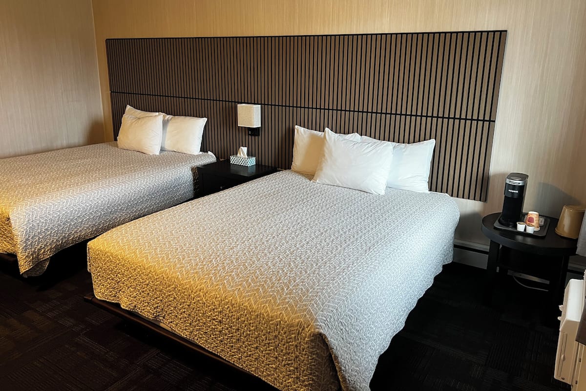 A hotel room with two beds, a nightstand with a coffee maker, and a wall-mounted lamp.