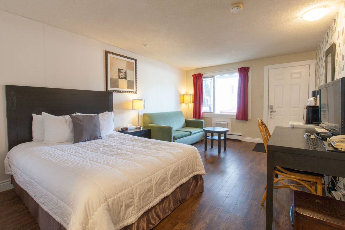 A hotel room with a large bed, a green armchair, a small table, and a desk with a computer and phone.