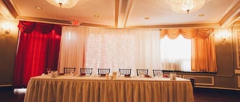 A banquet hall with a round table set for a meal, chandeliers, and curtains.