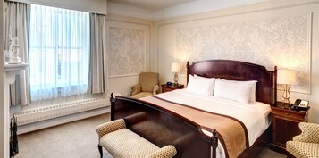 Habitación de hotel con una cama grande, dos mesitas de noche con lámparas, una silla, un panel decorativo en la pared y una ventana con cortinas.