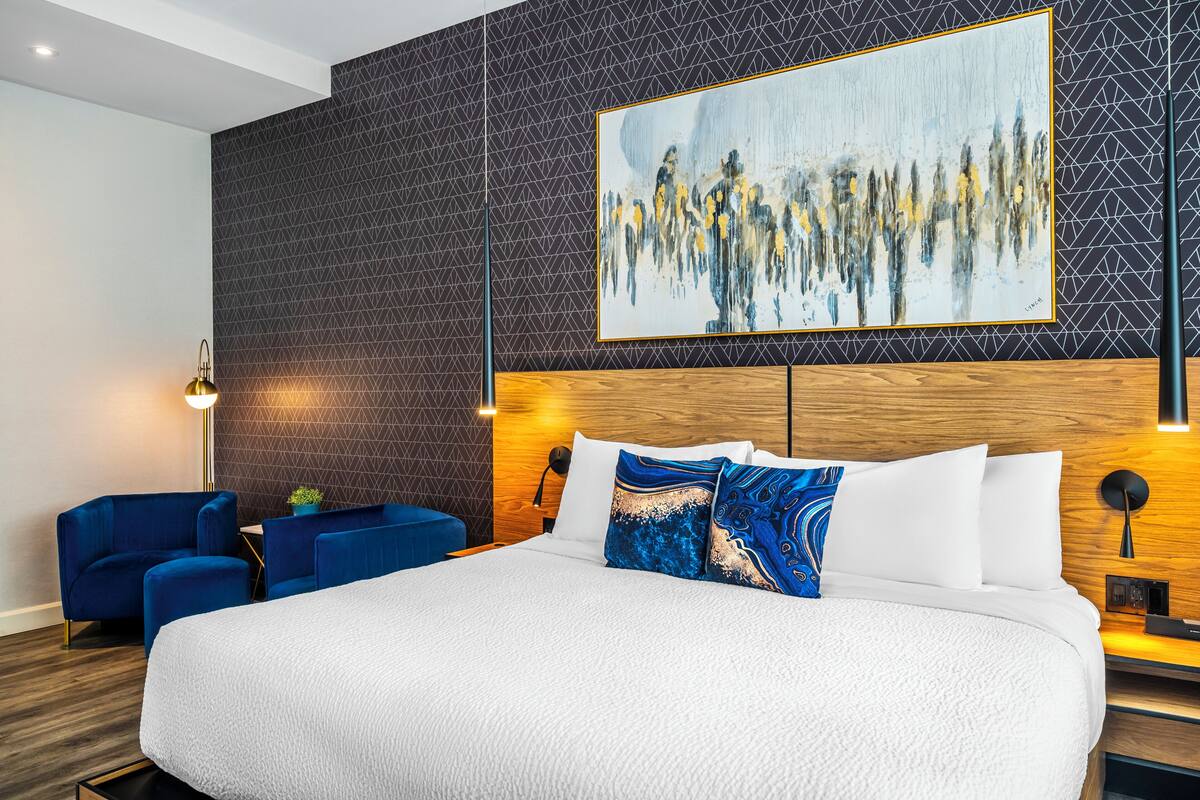 A hotel room with a large bed, a blue armchair, a small table, and a painting on the wall.