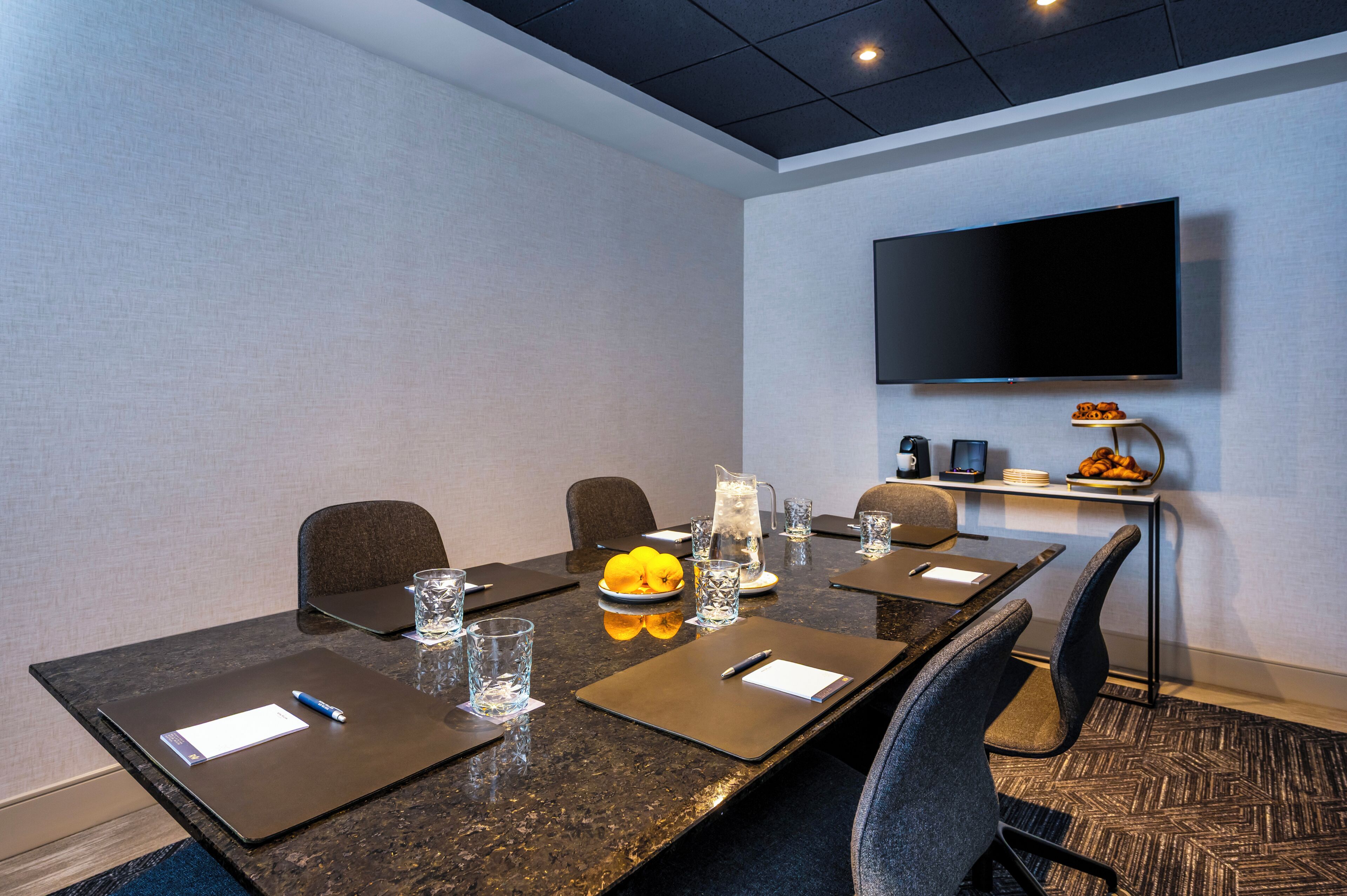 Un salón de conferencias con una larga mesa, sillas, un televisor montado en la pared y un pequeño estante con aperitivos.