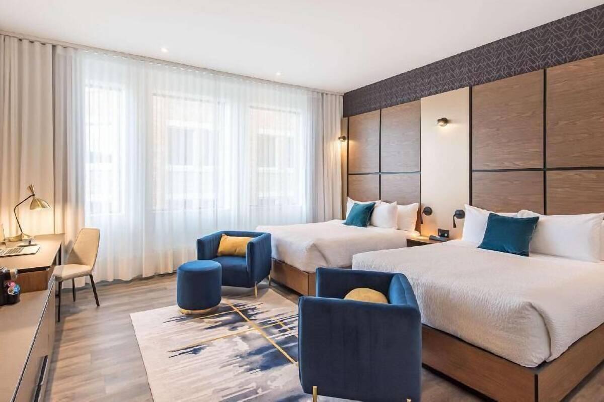 A hotel room with two beds, a desk, a chair, and a large window with sheer curtains.