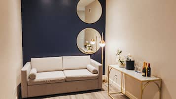 A room with a beige sofa, a round mirror, a small table with bottles, and a gold-framed console table.