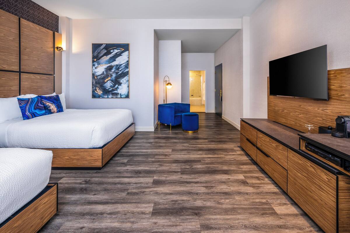 A modern hotel room with a large bed, a flat-screen TV, wooden flooring, and a hallway leading to a bathroom.