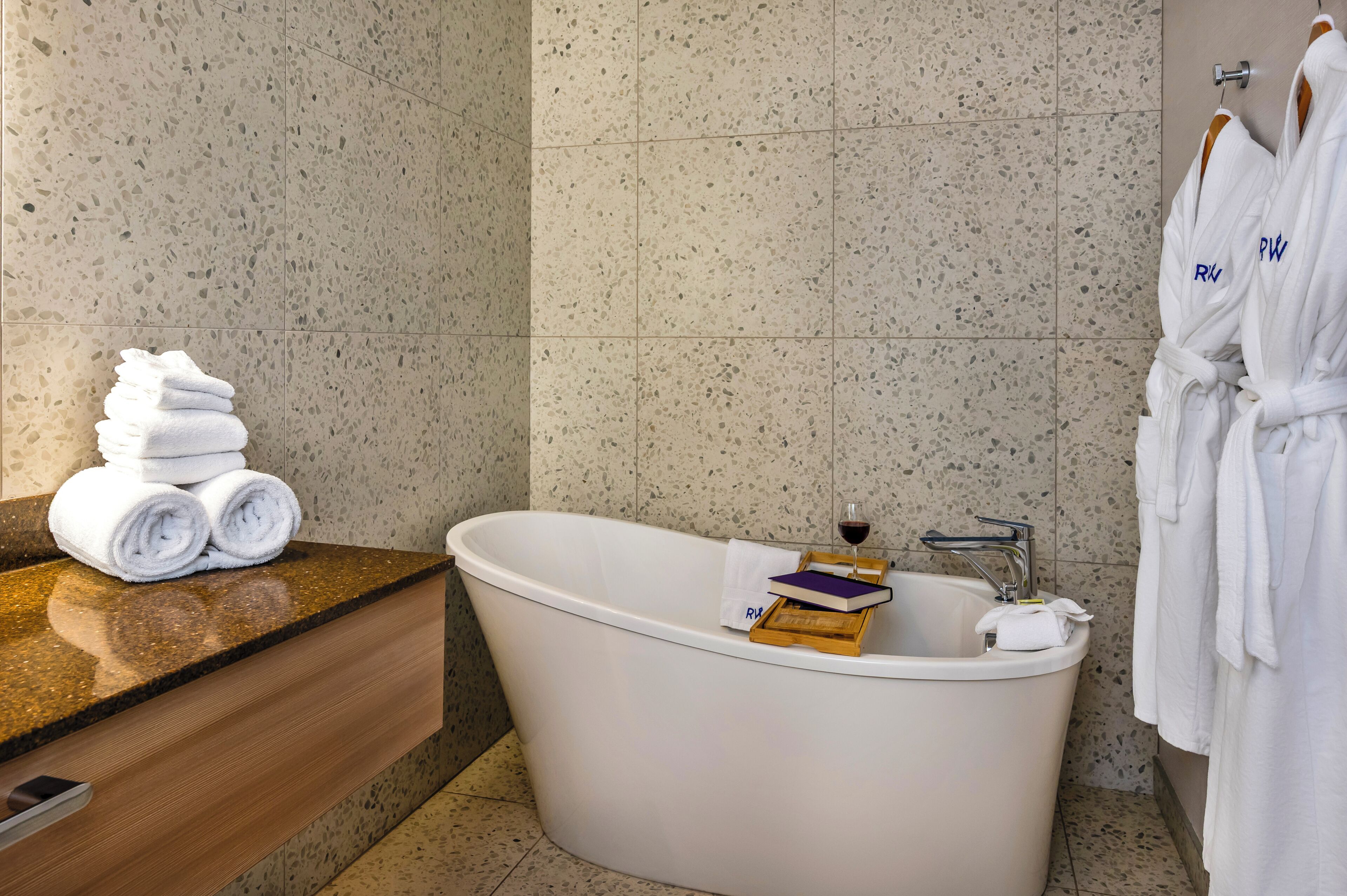 Ein modernes Badezimmer mit einer freistehenden Badewanne, einem Stapel gefalteter Handtücher und zwei weißen Bademänteln, die auf einem Gestell hängen.