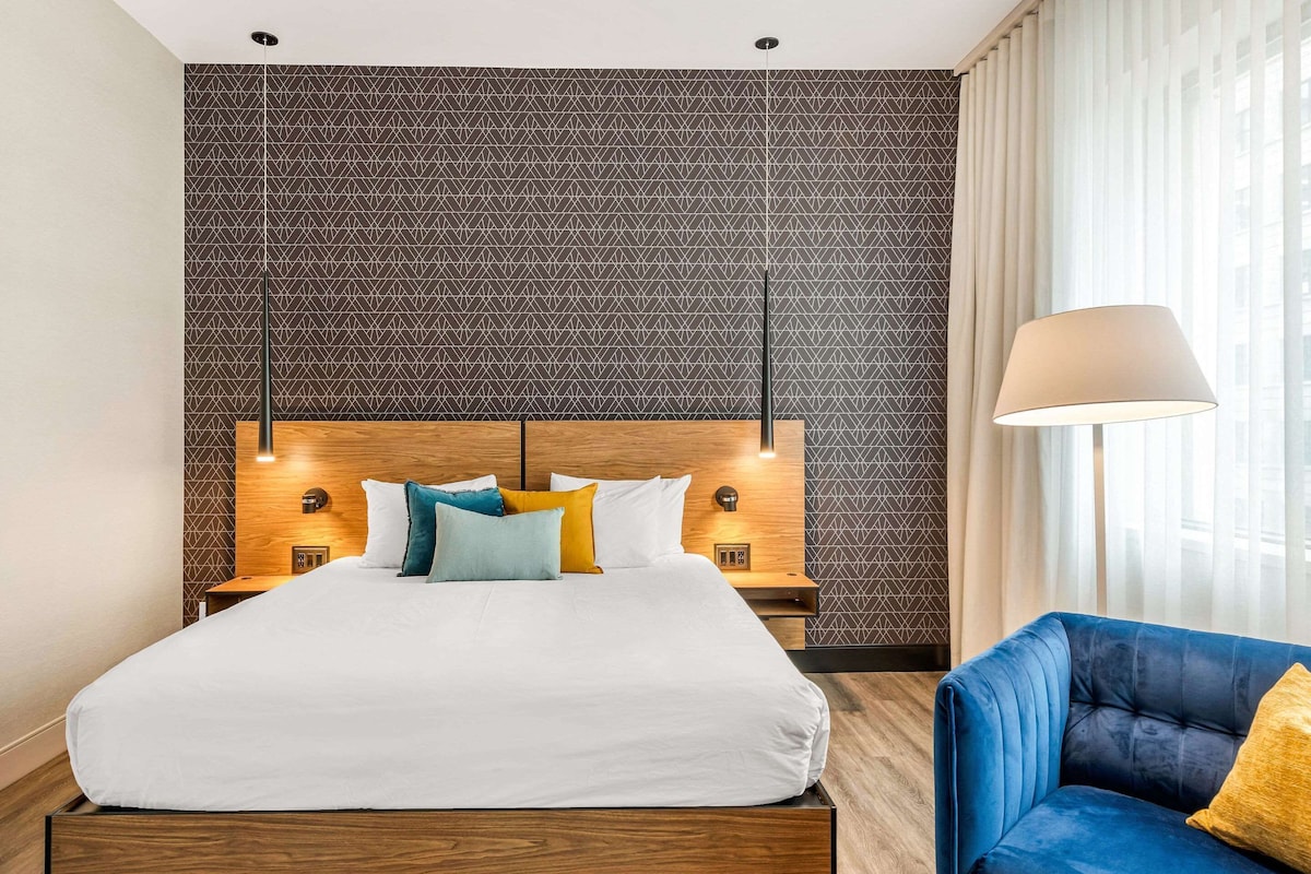 A hotel room with a large bed, a blue armchair, a lamp, and a patterned wall.