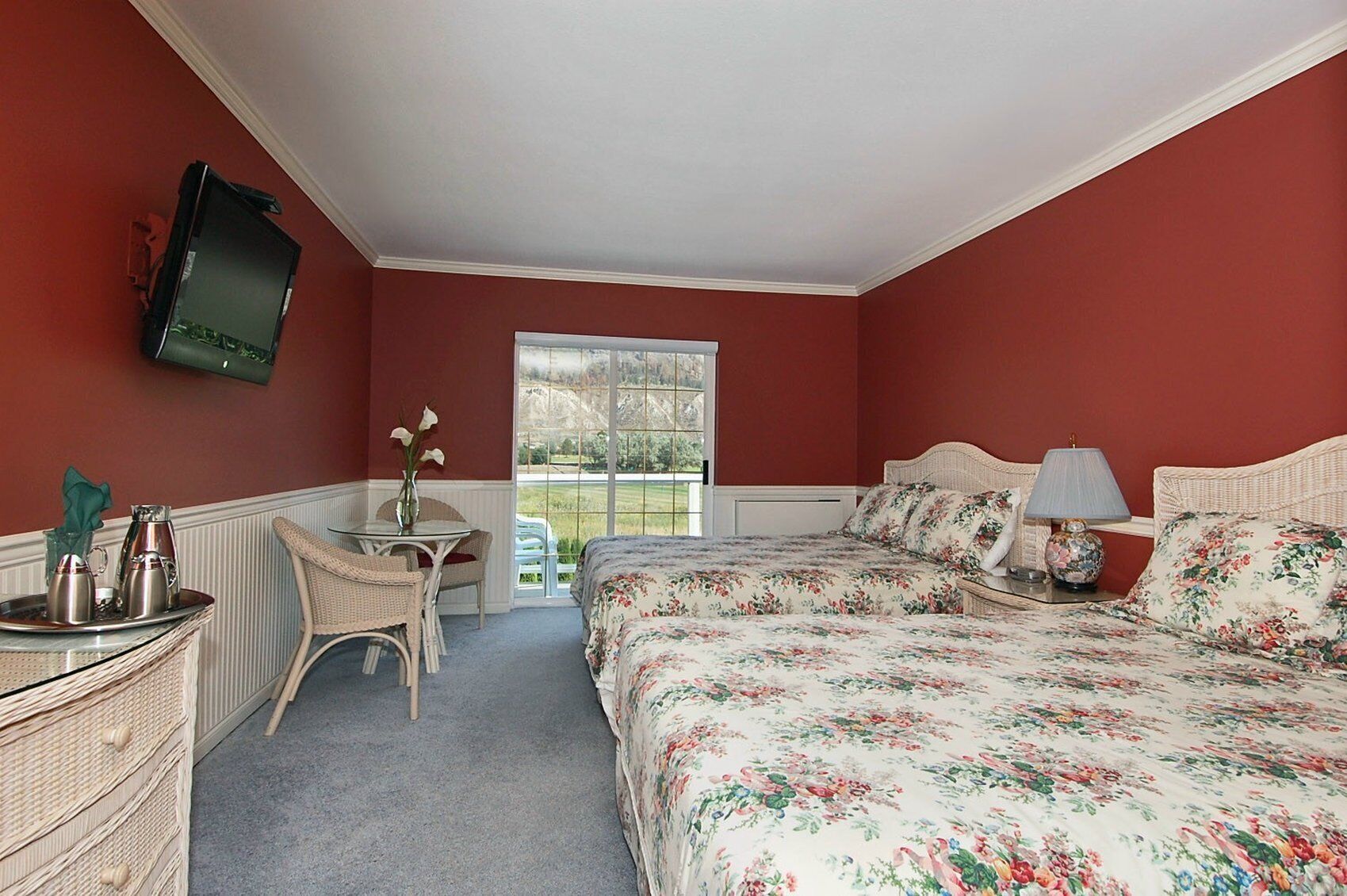 Ein Schlafzimmer mit Bett, Nachttisch mit Lampe, Stuhl, kleinem Tisch mit Blumenvase und Fenster mit Blick ins Grüne.
