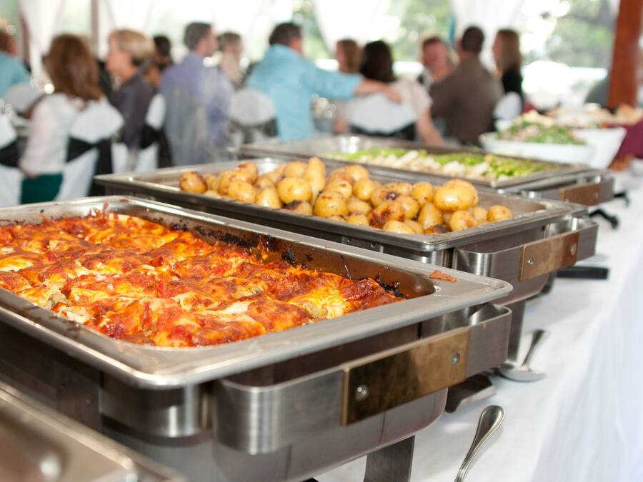 Buffet mit verschiedenen Speisen, darunter eine große Lasagneplatte, eine Platte mit Bratkartoffeln und eine Platte mit gemischtem Gemüse.