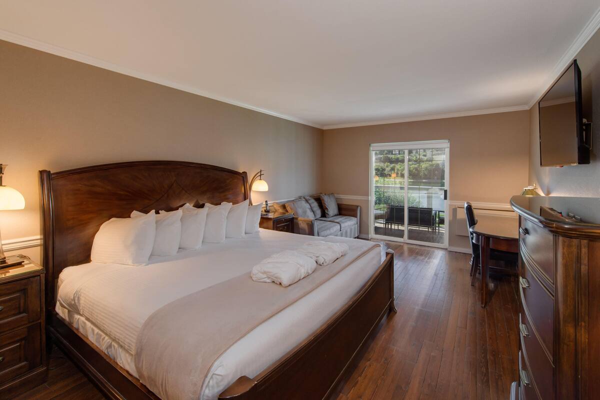 A bedroom with a large bed, a desk, and a chair. There is a window with a view of greenery outside.