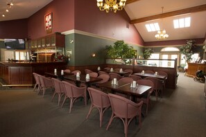 A restaurant with high ceilings, chandeliers, and a bar area. Tables are set with candles and chairs are upholstered in a pinkish fabric.