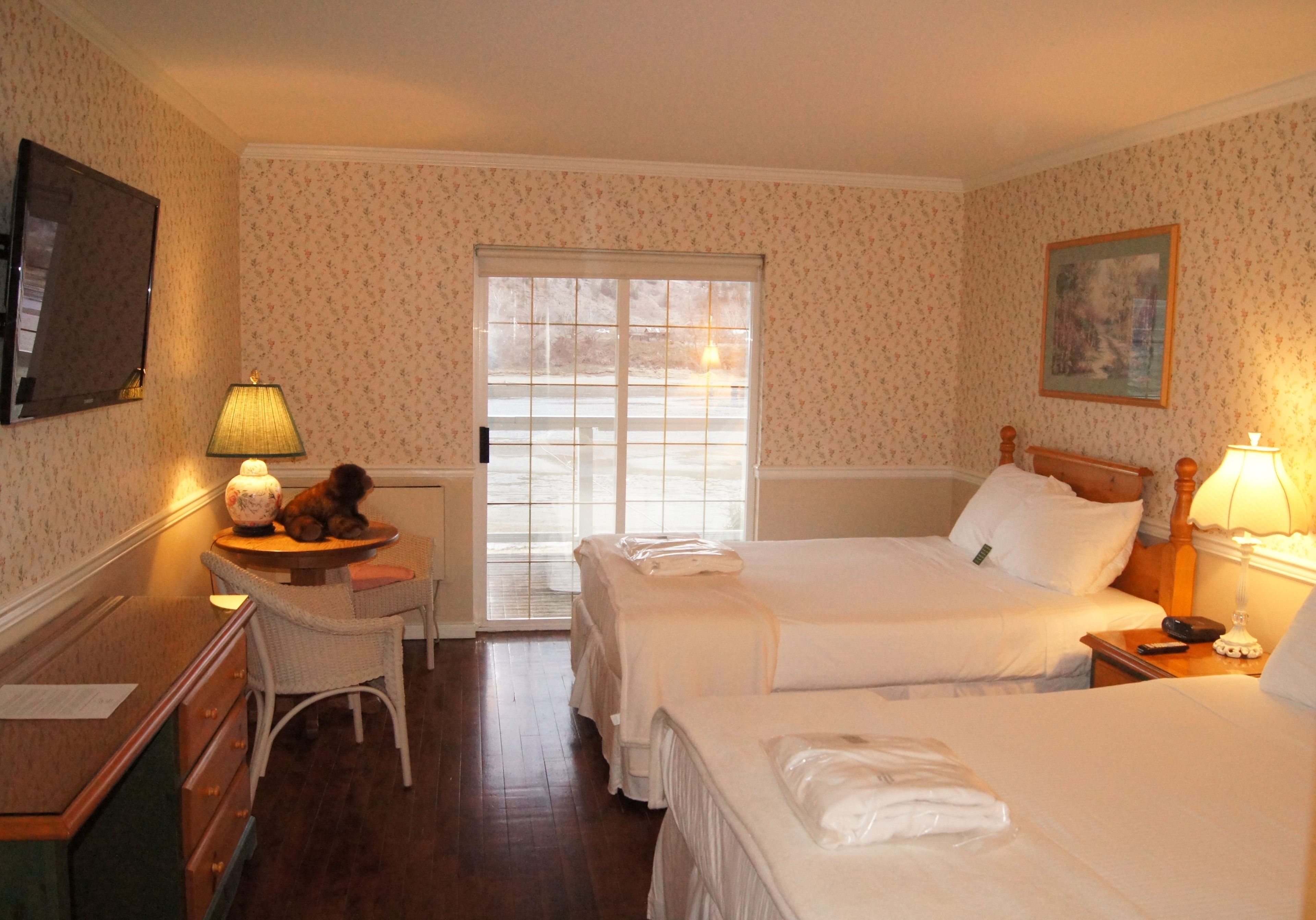 A hotel room with two beds, a desk, a chair, a lamp, and a window with a view of the outside.