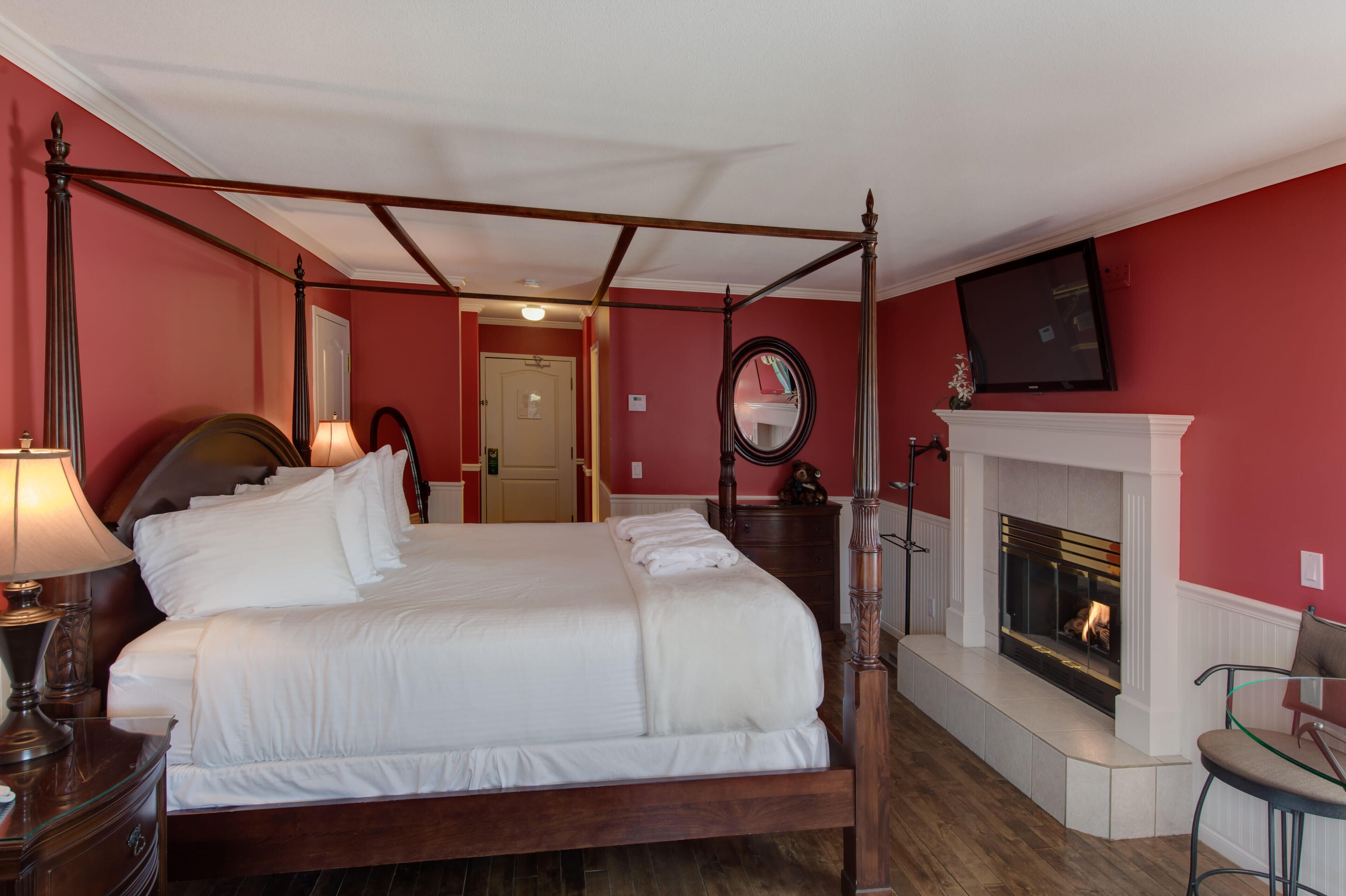 A four-poster bed with white linens, a wooden headboard, and a bedside table with a lamp. A fireplace with a white mantel is visible in the background.
