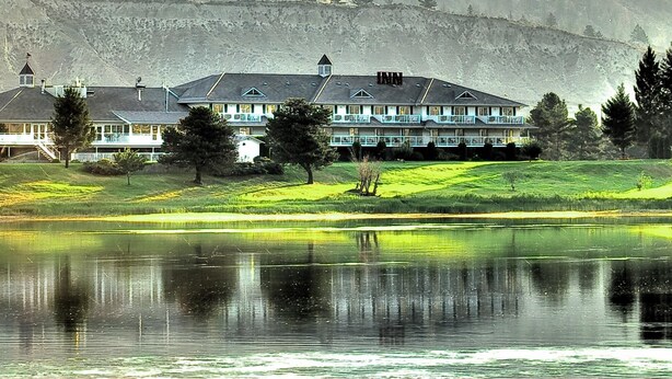 A large, multi-story hotel with a prominent roof and multiple balconies, situated beside a calm lake with reflections. - South Thompson Inn & Conference Centre (Kamloops)