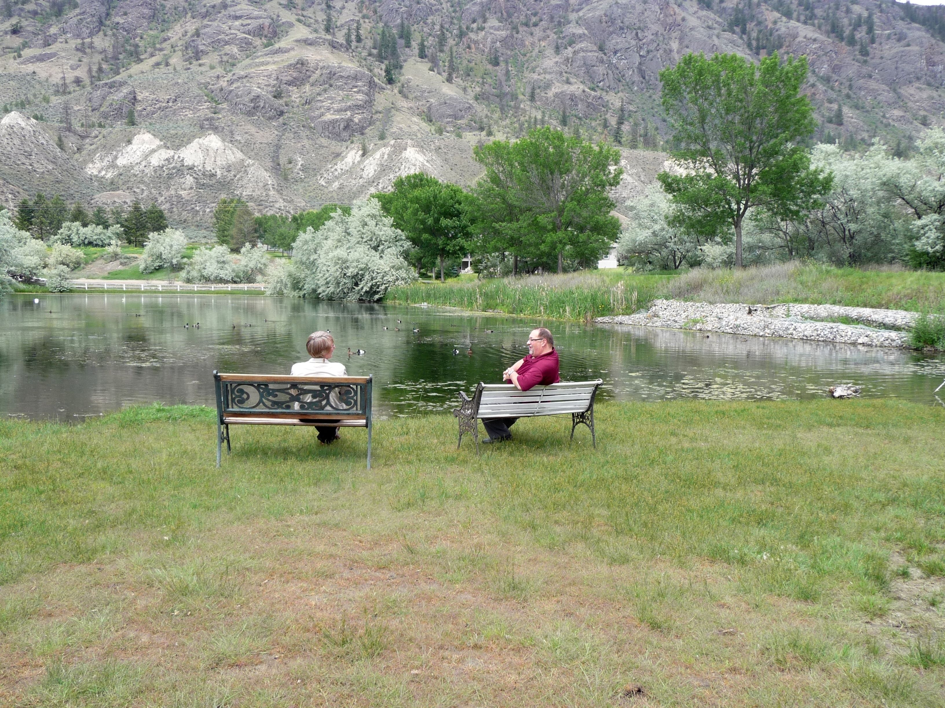 Dos bancos mirando a un lago con patos, rodeado de hierba y árboles, con montañas al fondo.