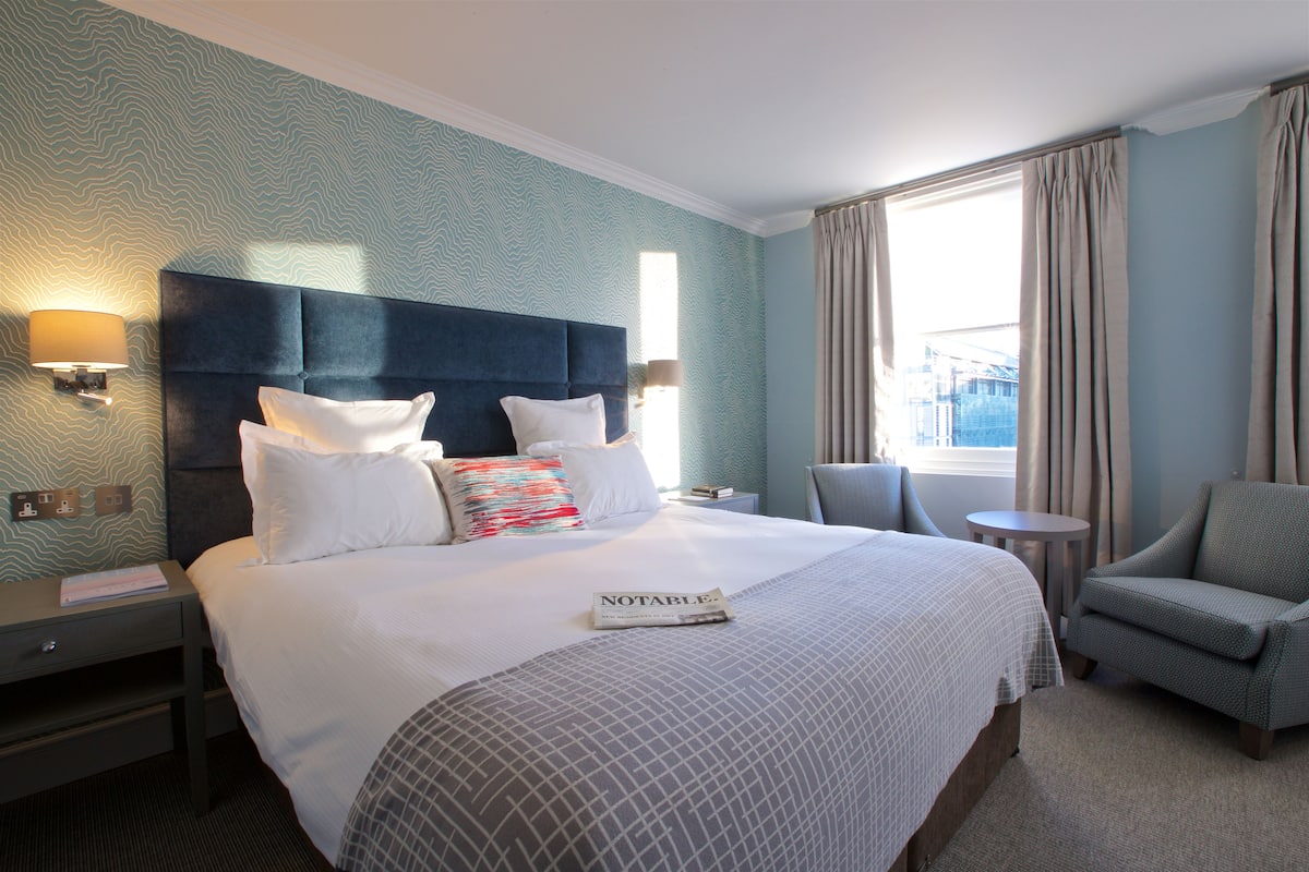Habitación de hotel con una cama grande, dos lámparas de noche, una mesita, una silla y un ventanal con cortinas.