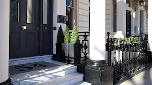Une porte noire portant le numéro 54, une porte blanche avec une fenêtre, et une clôture en fer noir agrémentée d’une jardinière.