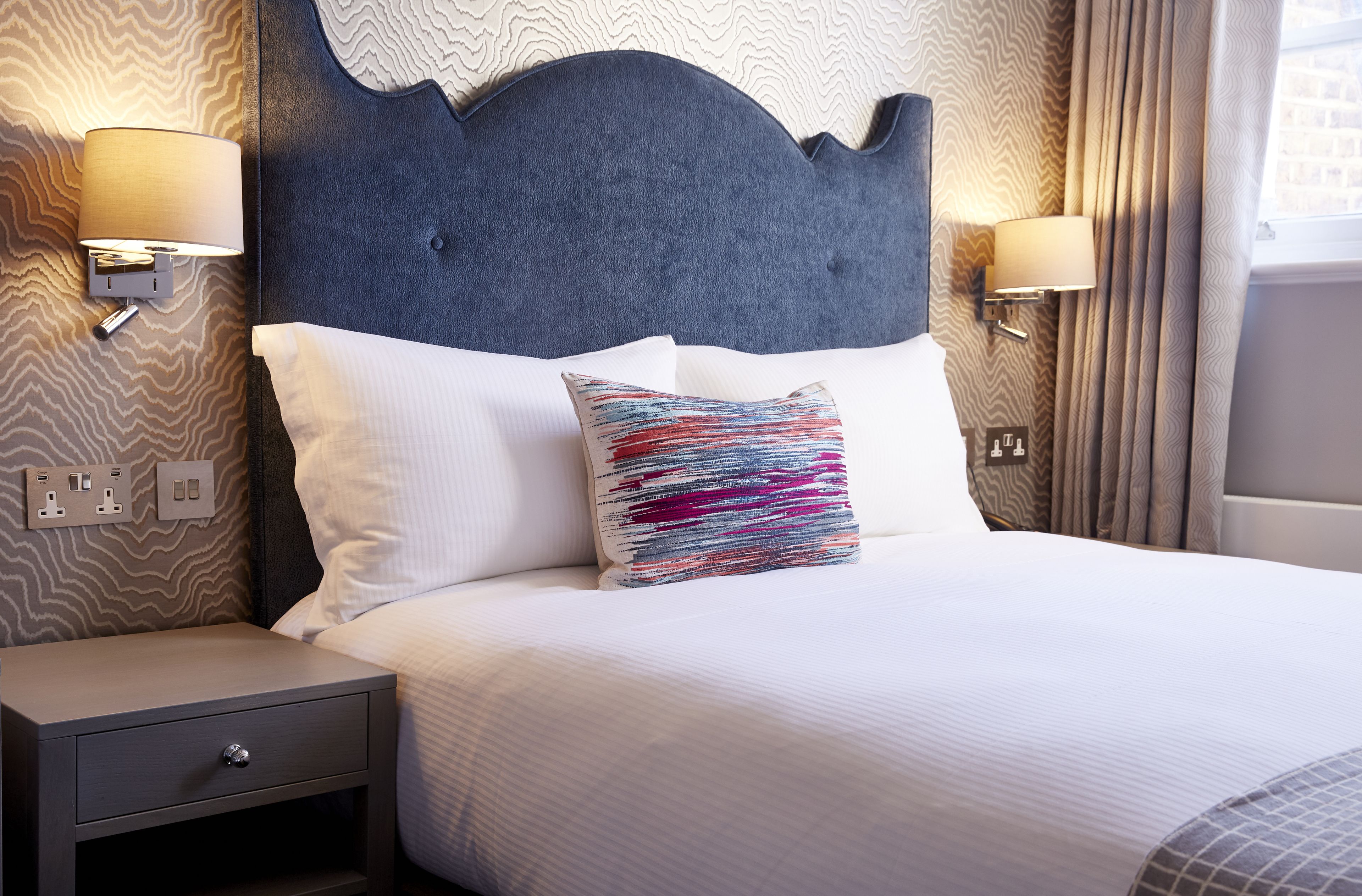 A neatly made bed with white linens, a decorative pillow, and a headboard with wall-mounted lamps.