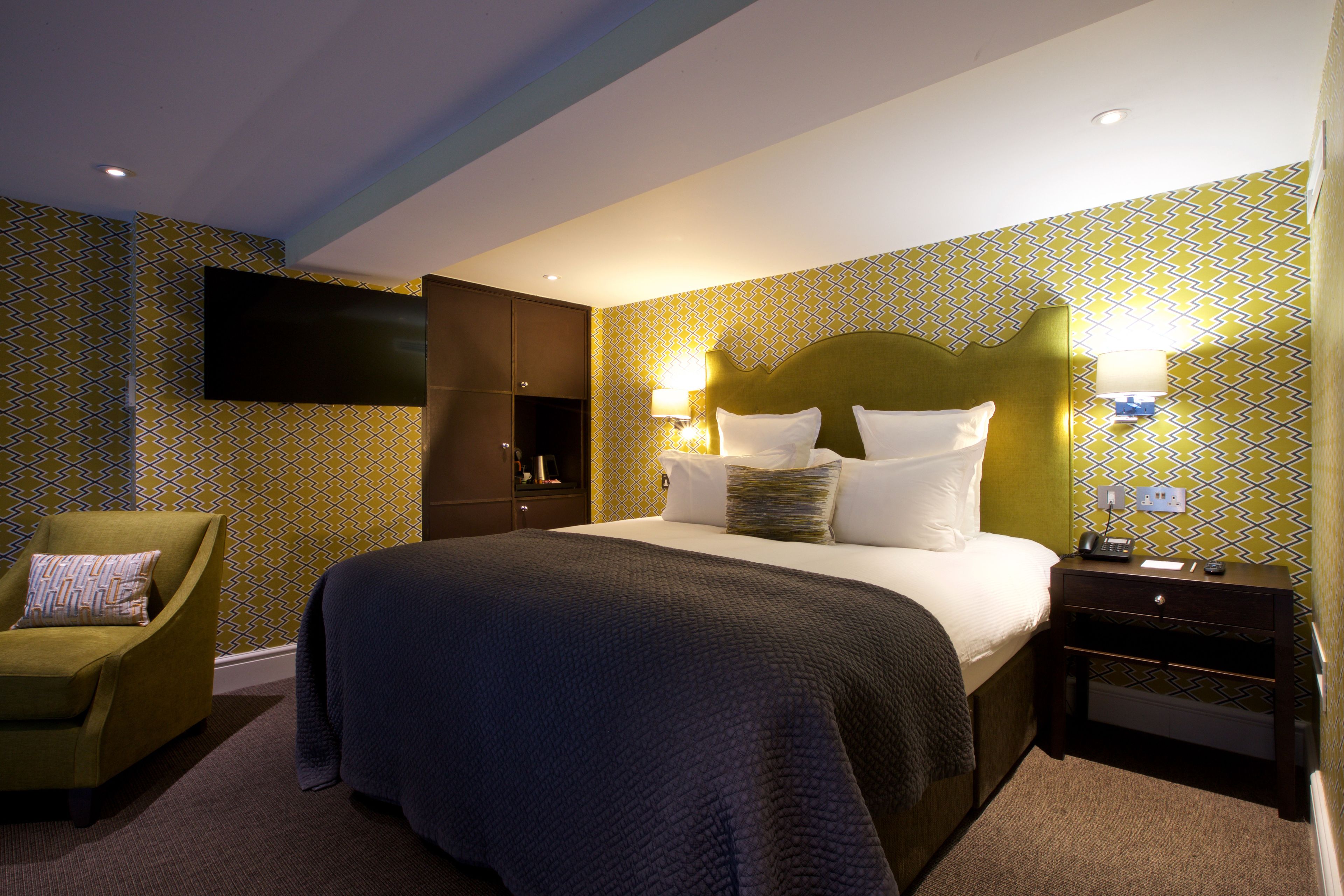 Une chambre d’hôtel avec un grand lit, un fauteuil vert, une table de chevet avec une lampe et un téléviseur fixé au mur.