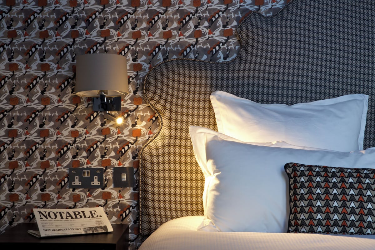 A neatly made bed with white linens, a patterned pillow, a wall-mounted lamp, and a small table with a magazine.