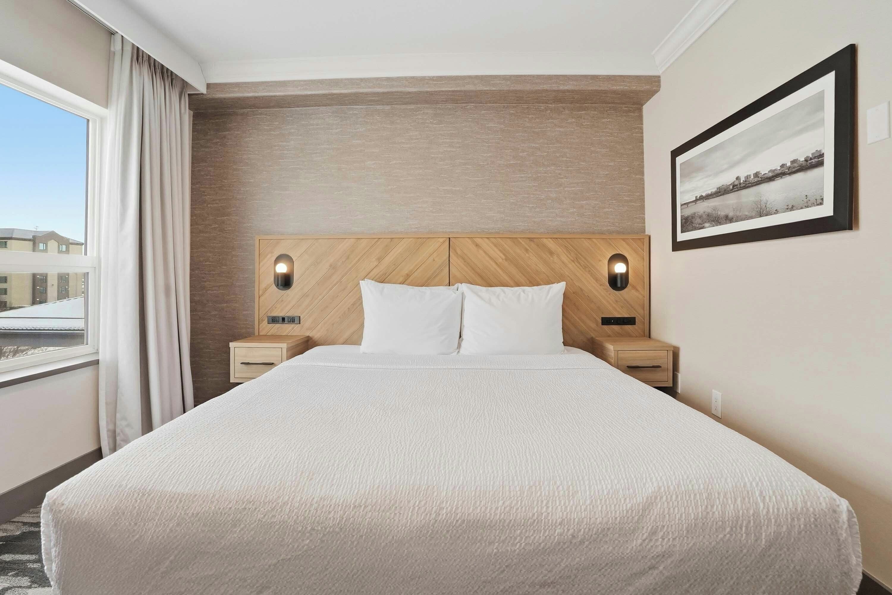 A hotel room with a large bed, wooden headboard, two bedside tables with lamps, a framed picture on the wall, and a view of buildings outside the window.