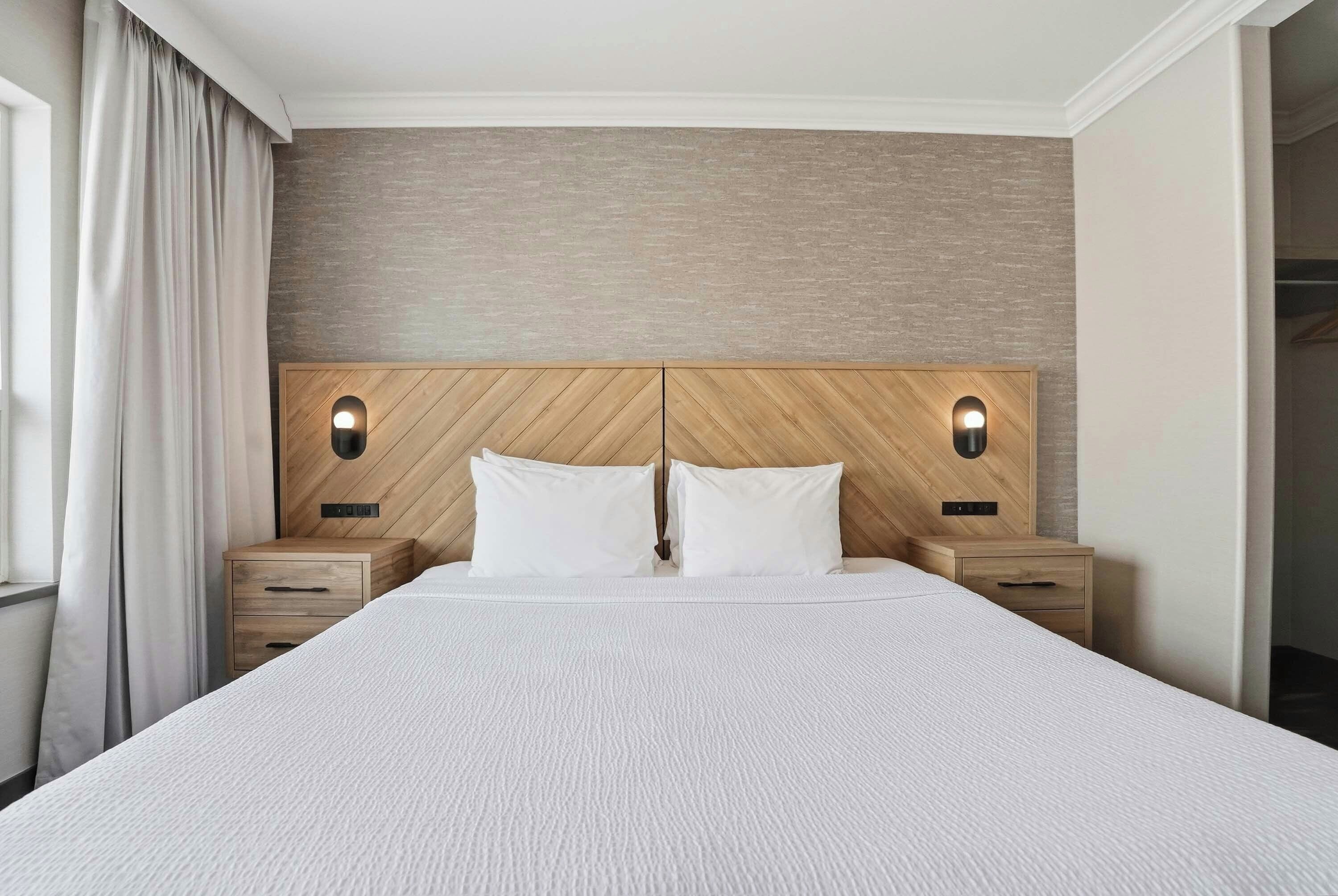 A hotel room with a large bed, wooden headboard, two bedside tables, and a view of the outside.