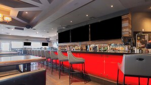 Un espace bar avec des tabourets rouges, un comptoir où sont exposées différentes bouteilles et un grand écran placé au-dessus du bar.