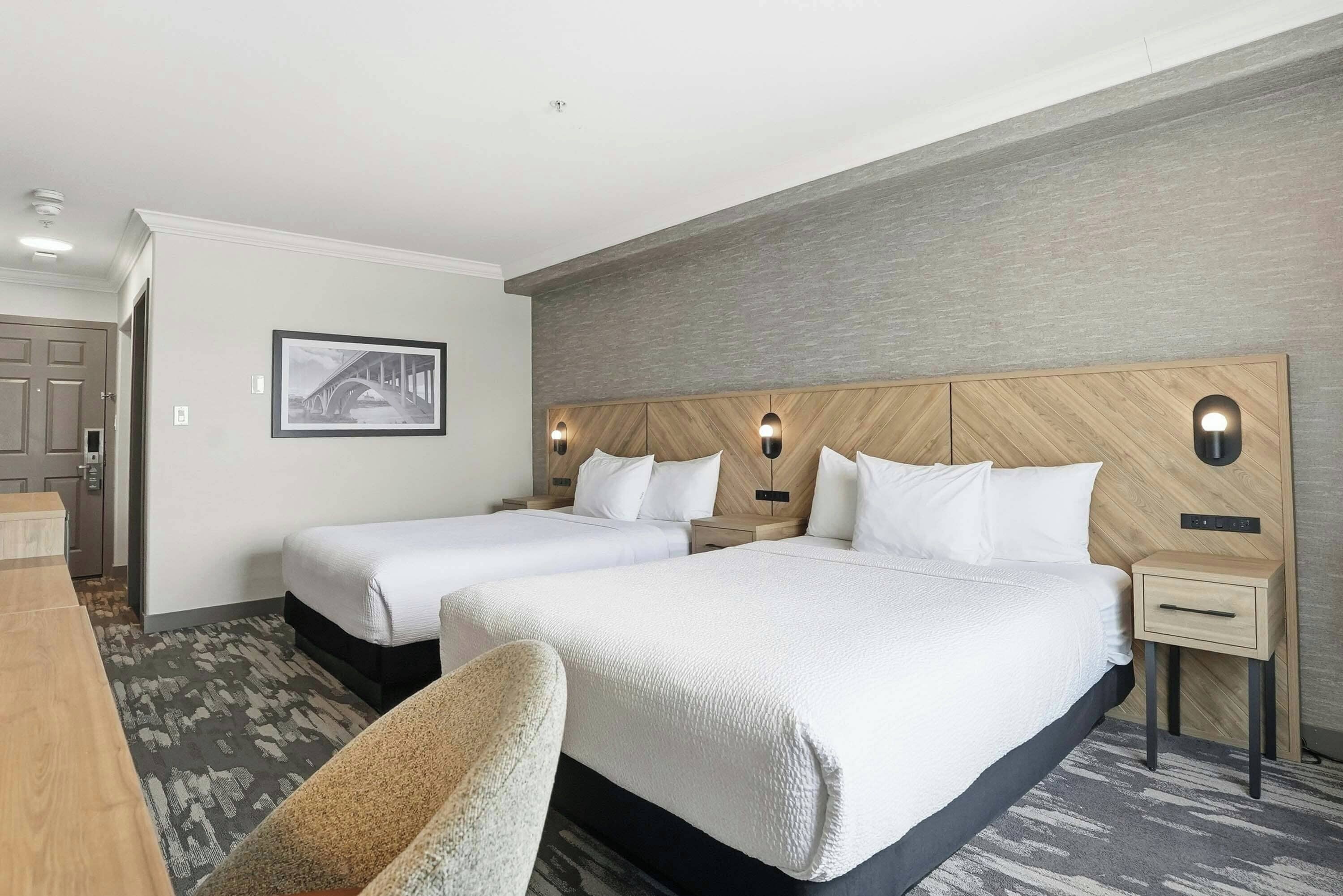 Une chambre d’hôtel avec deux lits, un bureau et une chaise. Il y a un tableau encadré au mur et une petite table avec une lampe.