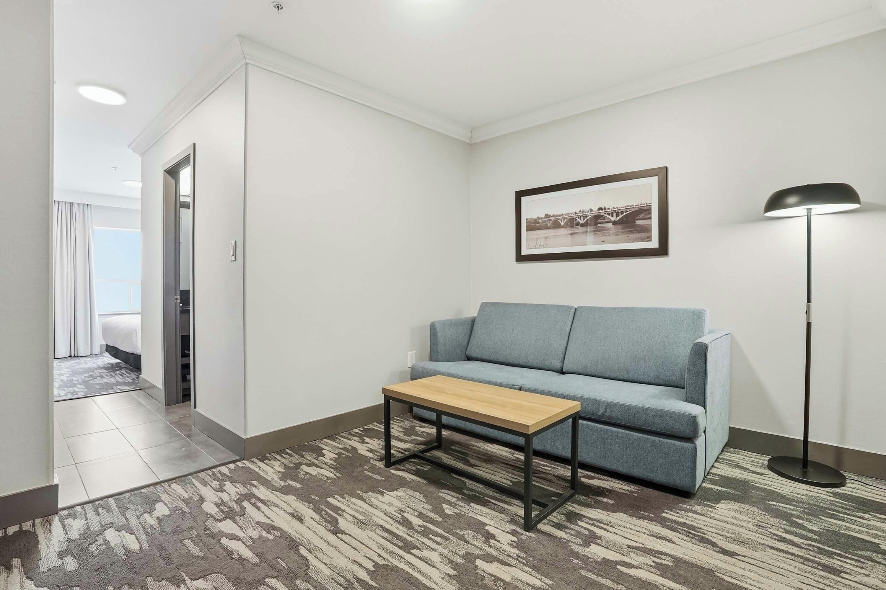 A modern hotel room with a grey sofa, a wooden coffee table, a framed picture on the wall, and a floor lamp.