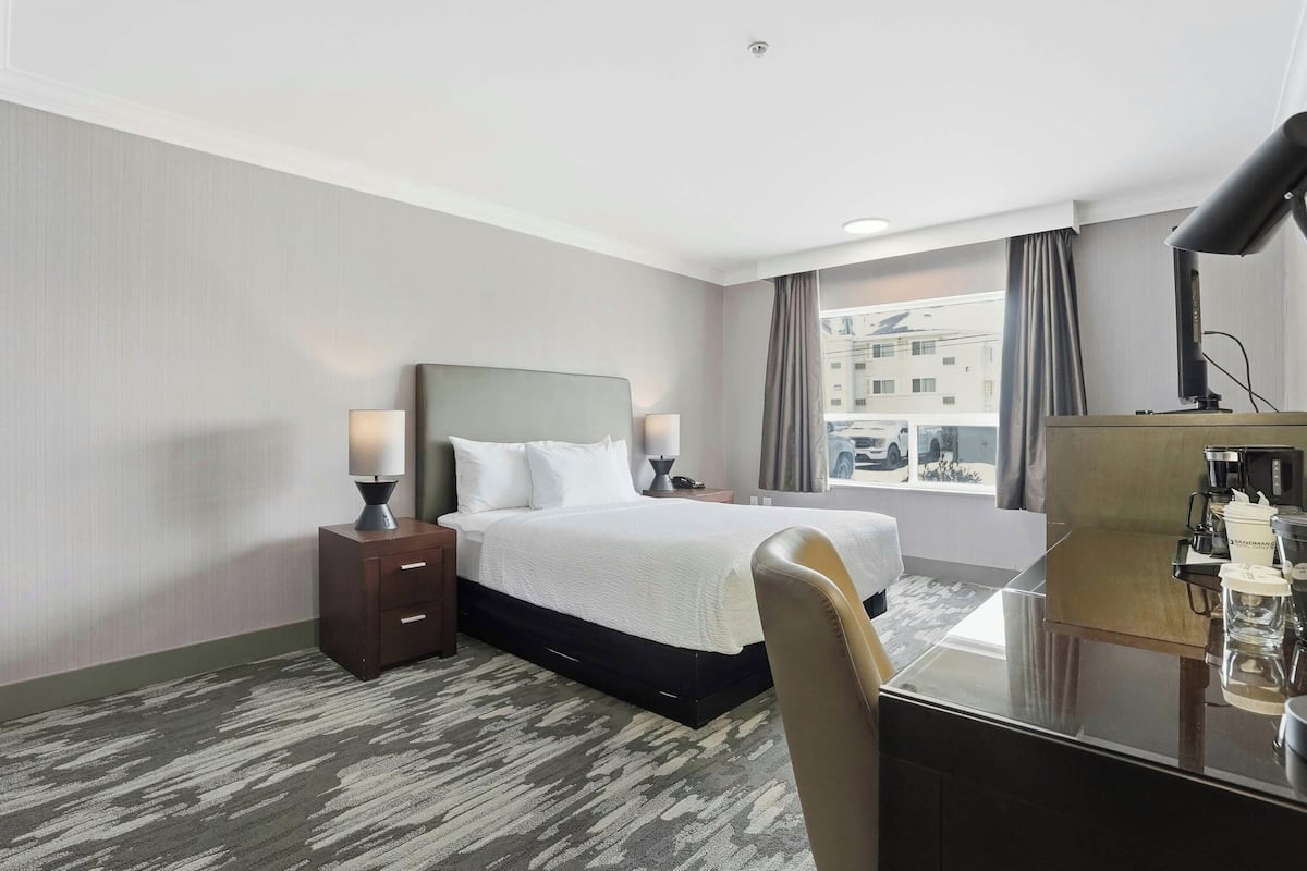 Une chambre d’hôtel comprenant un lit, des tables de chevet, un bureau, une chaise, une fenêtre avec des rideaux et une vue sur les bâtiments environnants.