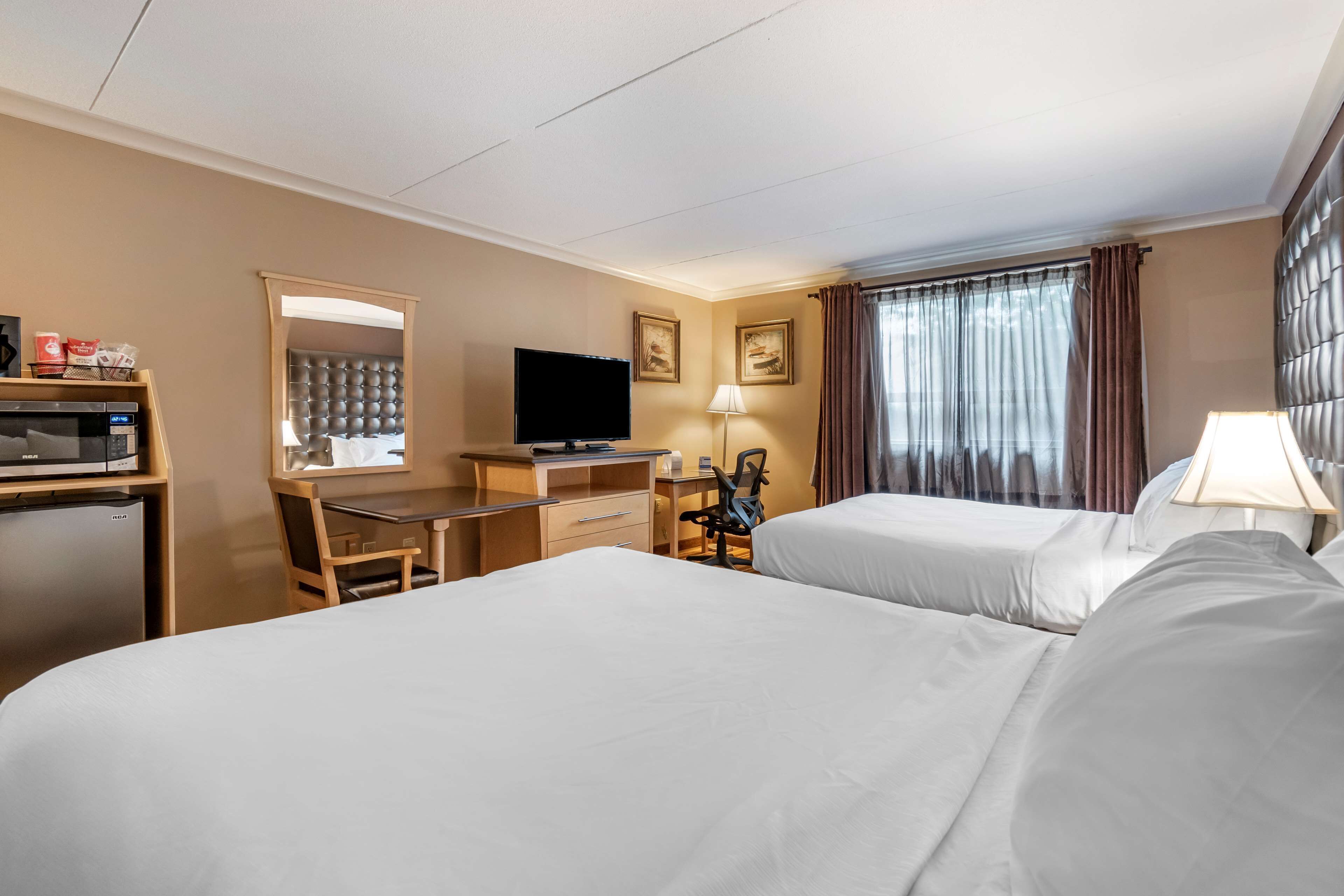 Ein Hotelzimmer mit zwei Betten, einem Schreibtisch mit Computer und Flachbildfernseher, einer Mikrowelle, einem kleinen Kühlschrank und einem Fenster mit Vorhängen.