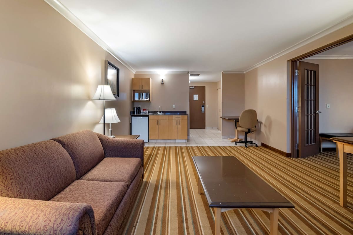 A hotel room with a sofa, a small table, a kitchen area, and a door leading to another room.