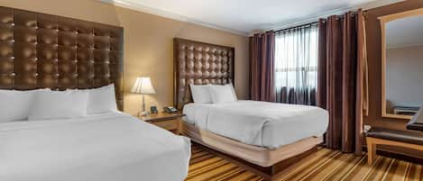 A hotel room with two beds, a large mirror, a bedside table with a lamp, and a striped carpet.