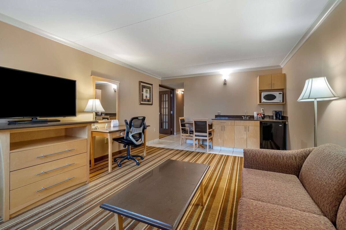 A hotel room with a flat-screen TV, a desk with a chair, a sofa, a kitchen area with a microwave, and a lamp.