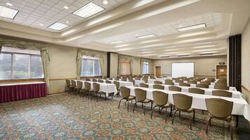 A conference room with rows of tables and chairs, large windows with curtains, a carpeted floor, and a whiteboard.