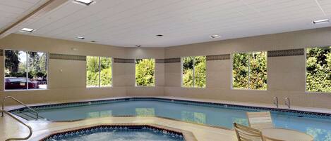 An indoor swimming pool with a hot tub, surrounded by windows with a view of greenery.