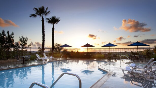 A swimming pool with lounge chairs and umbrellas, surrounded by palm trees and a sunset view.